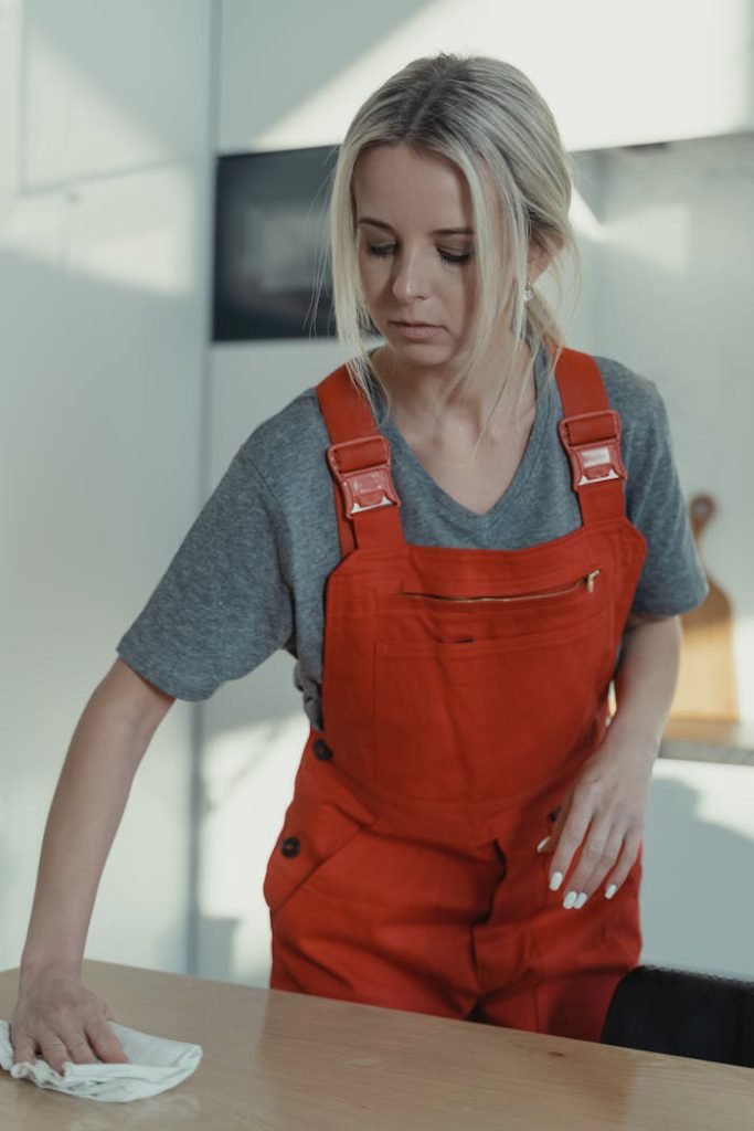 Blonde woman in red overalls sanitizing a wooden table indoors, showcasing home cleaning service.